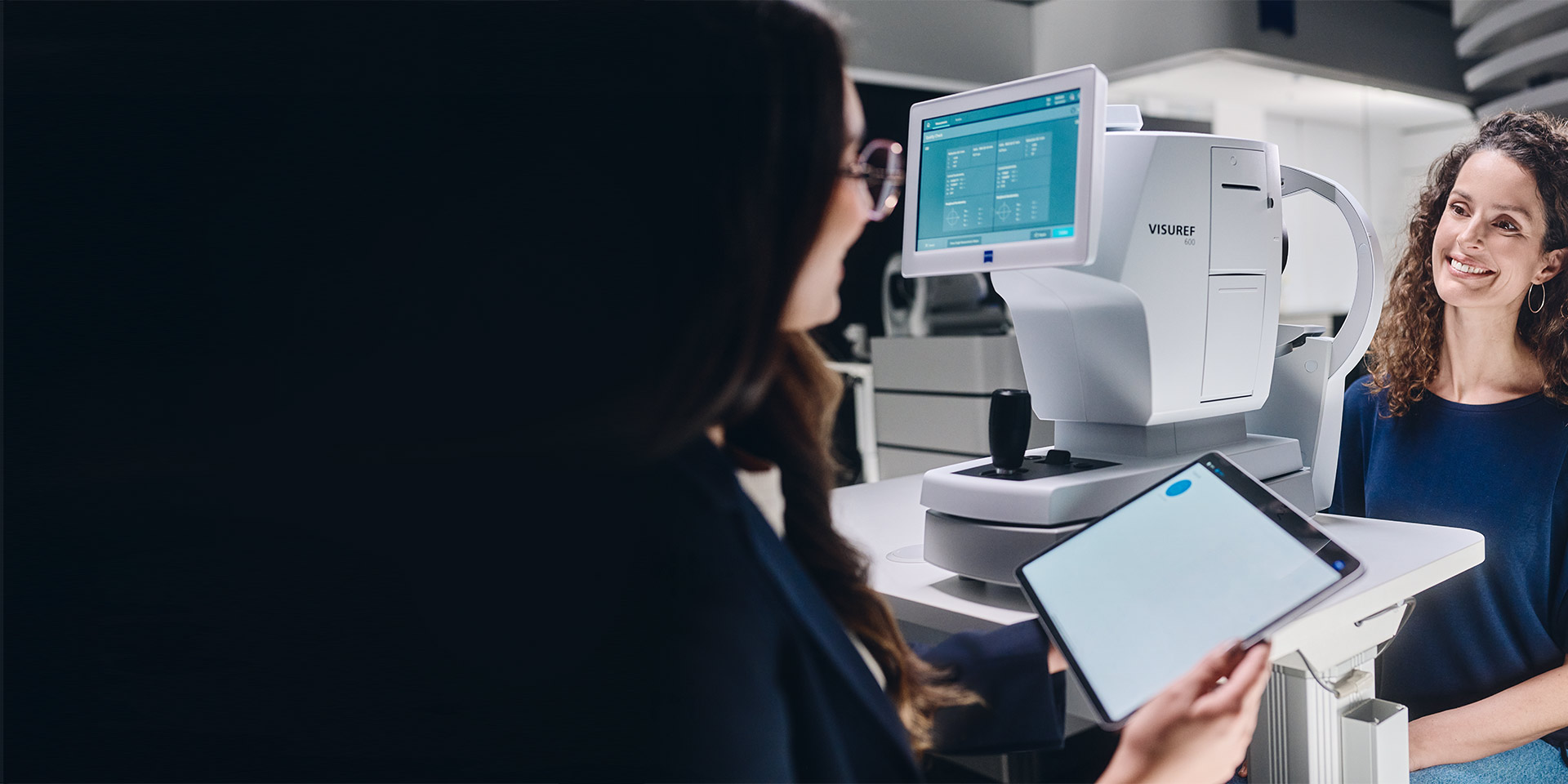 Naisoptikko katsoo tablettiaan istuen ZEISS VISUREF 600 -laitteen edessä ja puhuu asiakkaansa kanssa.