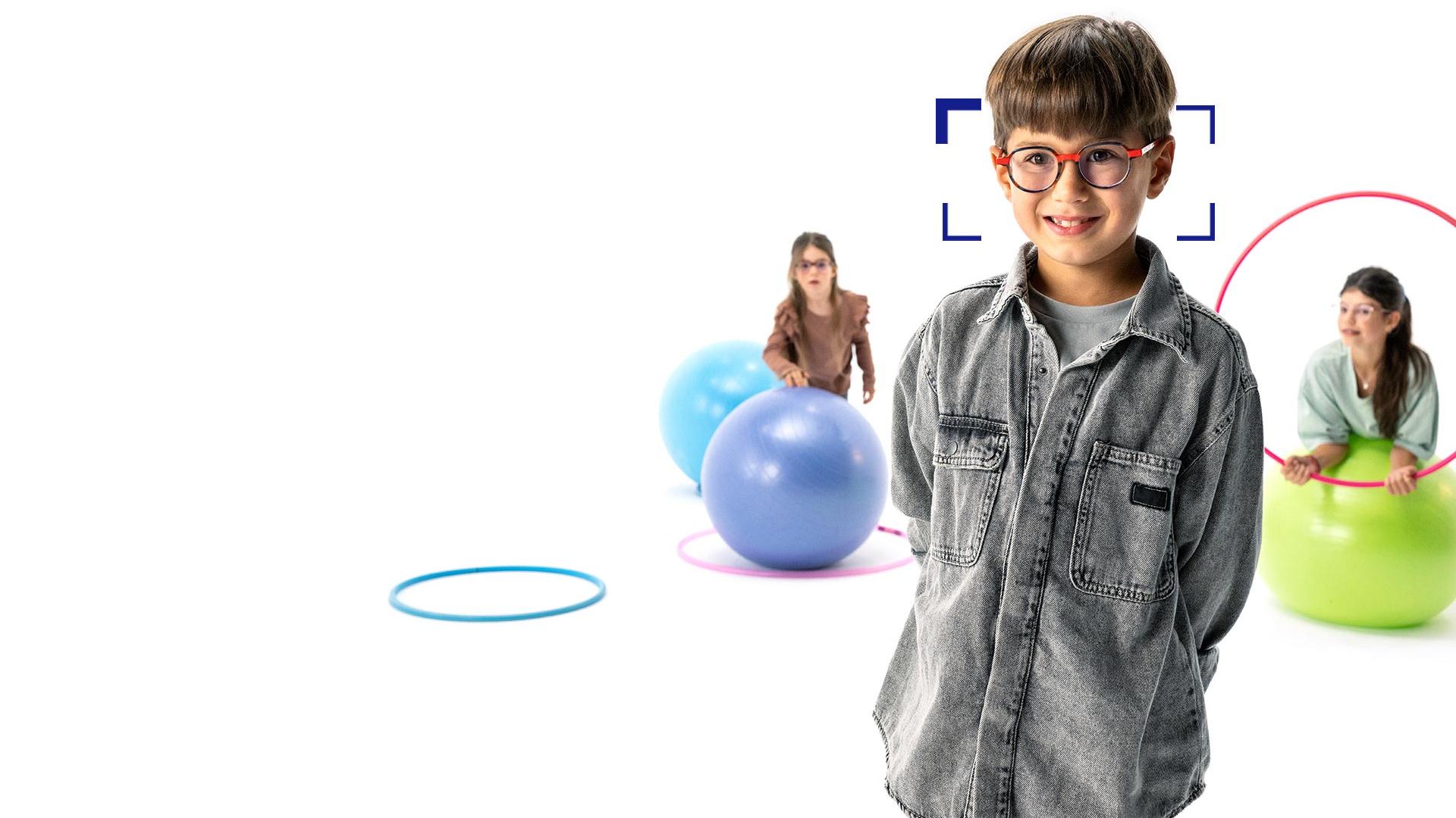 Ruskeahiuksinen poika silmillään ZEISS MyoCare -linssit seisoo etualalla ja hymyilee kameralle. Taustalla kaksi tyttöä päässään ZEISS MyoCare -linssit leikkivät vanteilla ja jumppapalloilla.