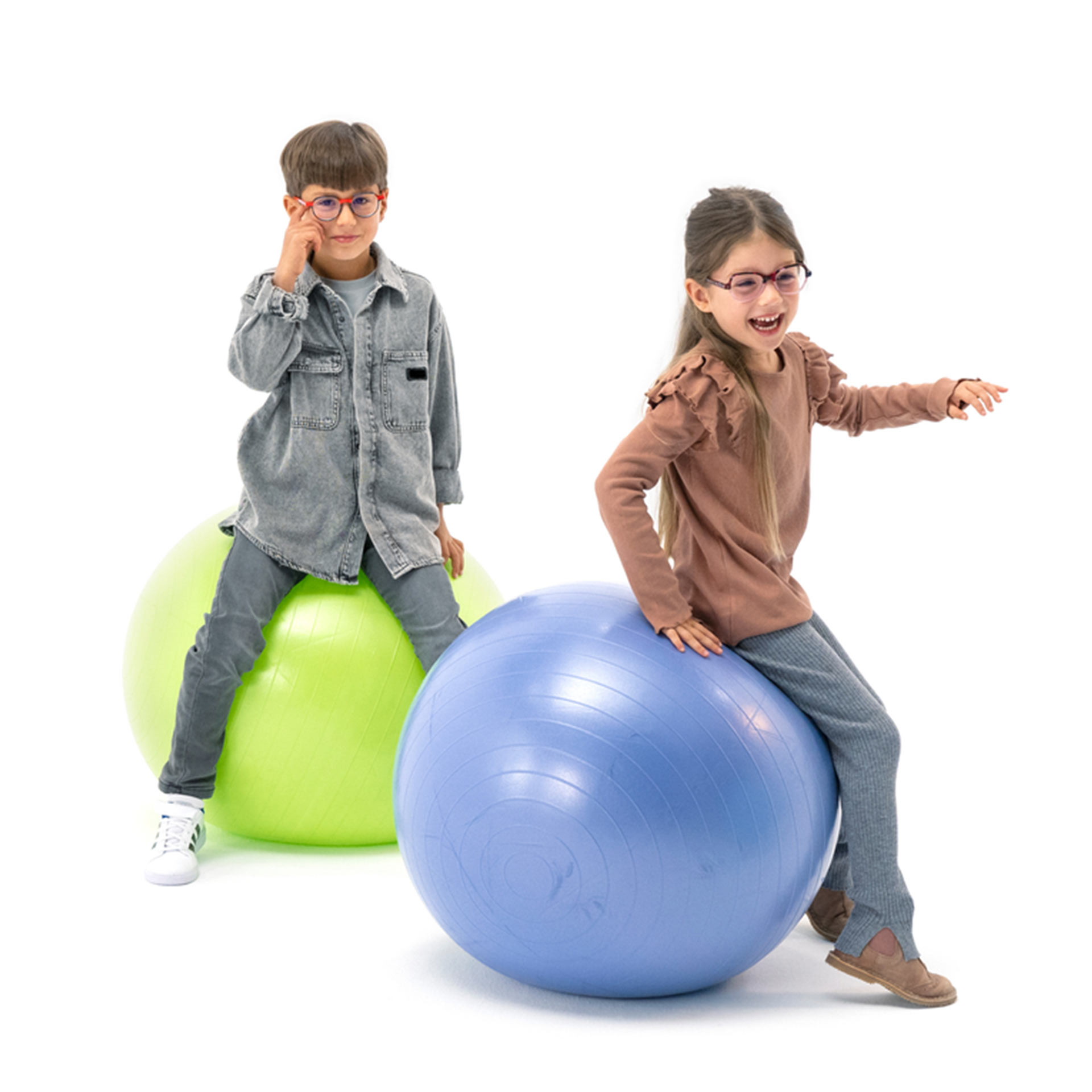 Poika ja tyttö, joilla molemmilla on silmälasit, pomppivat leikkisästi jumppapalloilla.