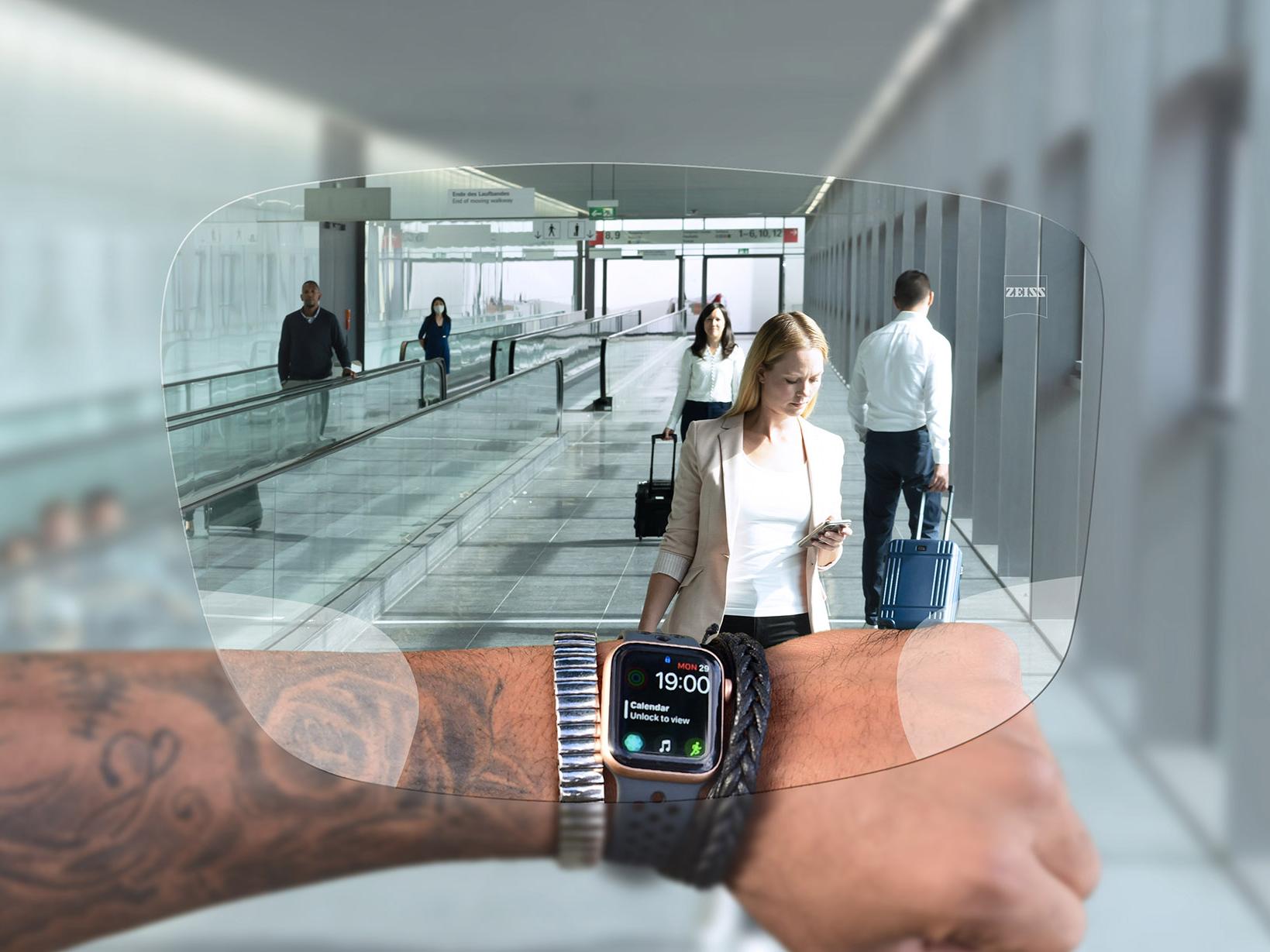 Henkilö katsoo kelloaan ZEISS SmartLife Digital -linssien läpi. Taustalla näkyy tyypillinen lentokenttätilanne, jossa ihmiset kävelevät liukuportaissa ja työntävät matkalaukkujaan. 
