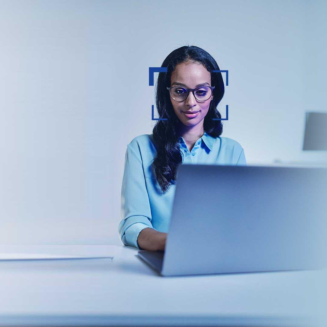 Nainen, jolla on mustat hiukset ja silmälasit, katsoo hymyillen kannettavaa tietokonetta