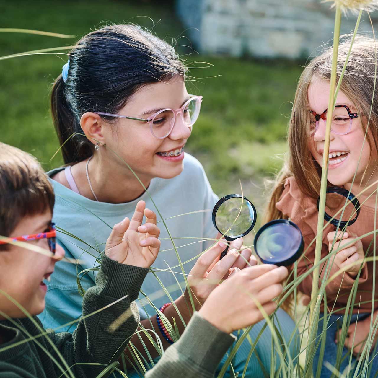 Ryhmä lapsia, joilla on silmälasit, leikkimässä suurennuslasien kanssa ja nauramassa ulkona