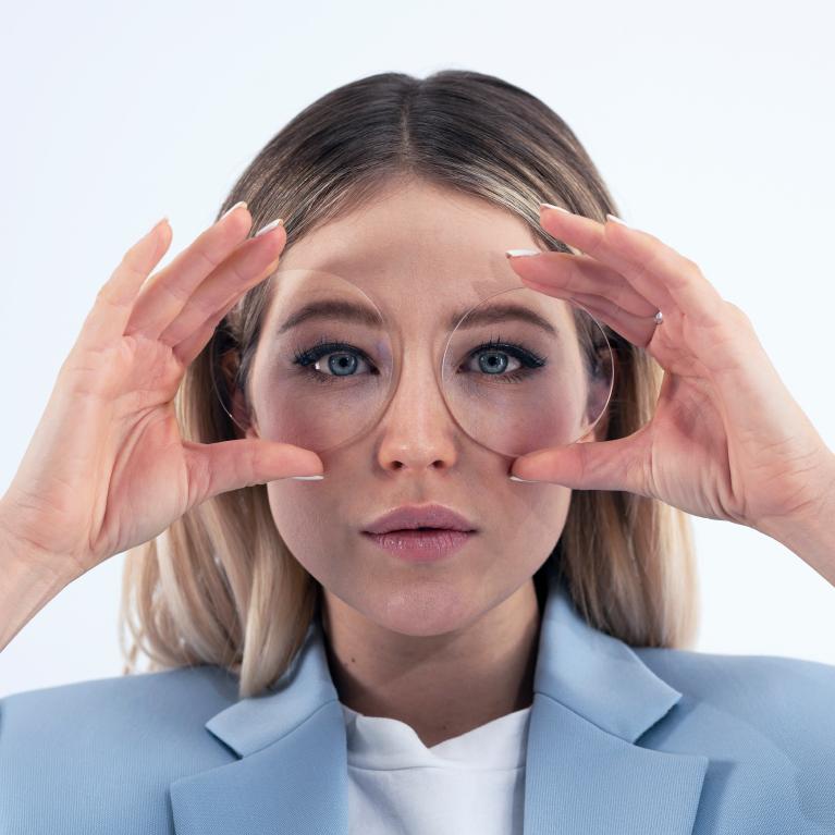 Nuori vaalea nainen pitää linssejä silmiensä edessä osoittaakseen, että näyttää hyvältä ilman hauskaa silmäefektiä, koska käyttää ZEISS ClearView -yksiteholinssejä.