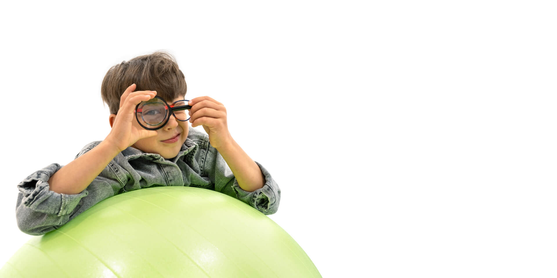 ZEISS Myopia Management -linssejä käyttävä poika nojaa jumppapalloon ja pitää suurennuslasia toisen silmänsä edessä.