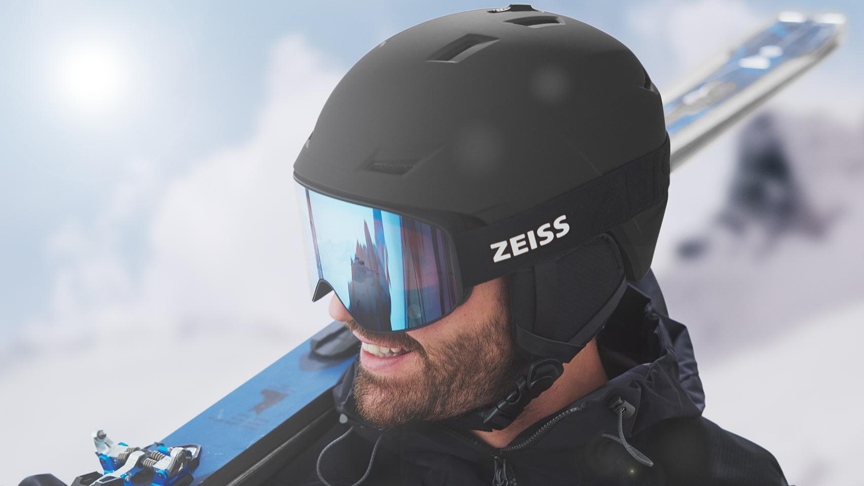 Miehellä on musta kypärä ja siniset peilipinnoitetut ZEISS-laskettelulasit ja olallaan sukset.